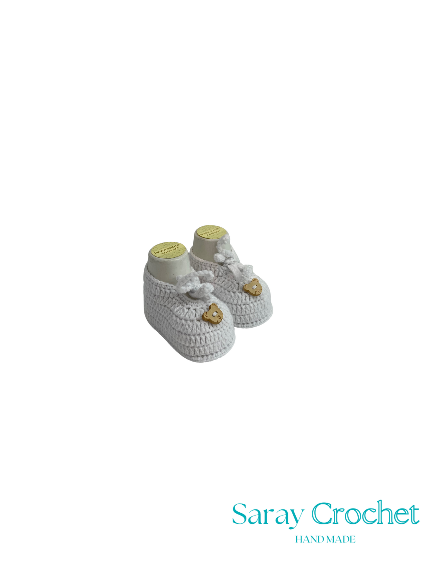 Pair of white baby booties with gold accents on a white background, branded 'Saray Crochet'.