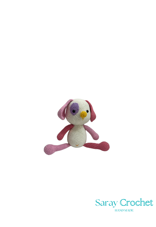 A crochet toy resembling a stylized dog with a pink body, white face, and purple eyes, with the brand name 'Saray Crochet' visible.
