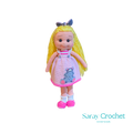 Handmade crochet doll with blonde hair, wearing a pink dress and white and pink shoes, standing against a black background.