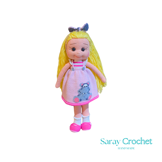Handmade crochet doll with blonde hair, wearing a pink dress and white and pink shoes, standing against a black background.