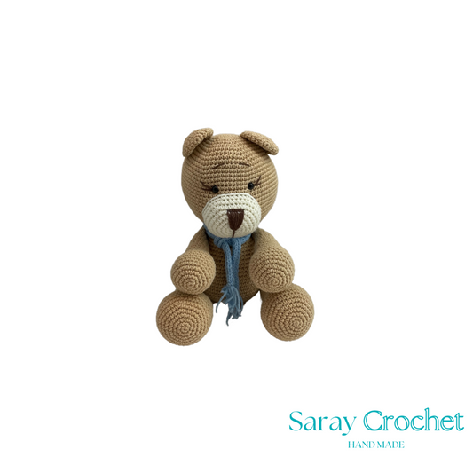 Crocheted teddy bear with a blue scarf on a white background, branded 'Saray Crochet'.