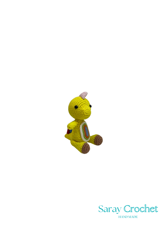 A small yellow plush toy dinosaur with a patterned fabric body, displayed against a white background with the text 'Saray Crochet' at the bottom.