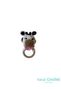 A crochet teething ring toy designed to look like a cow with a predominantly white body and black and brown details, attached to a wooden ring.