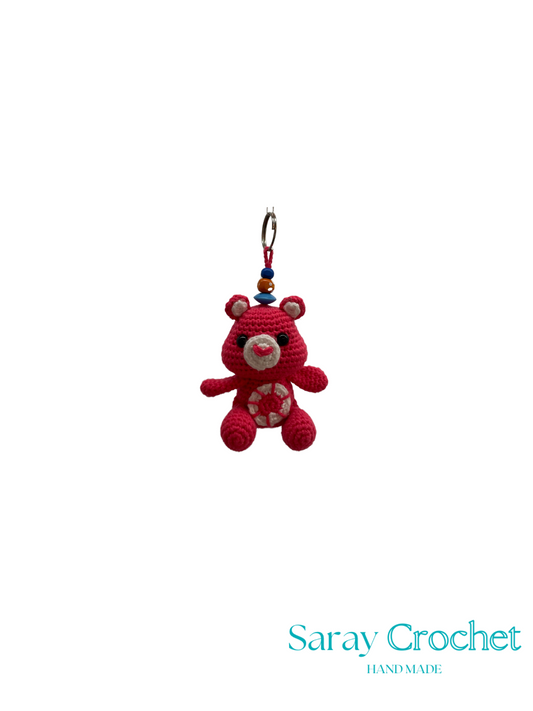 Red crochet teddy bear keychain with a white heart on a white background, branded 'Saray Crochet'.