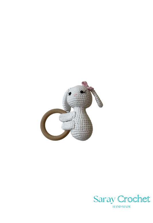 A crochet toy in the shape of a white rabbit with a pink flower on its head and a wooden ring attached to its body.