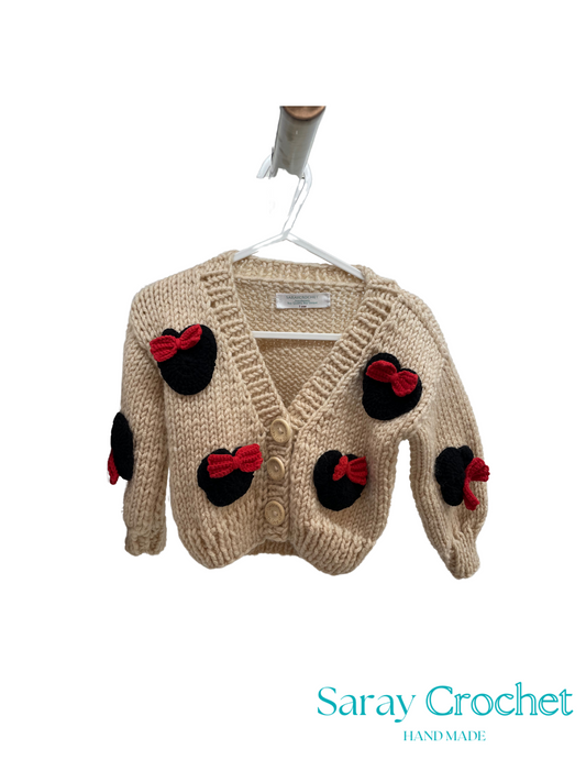 Beige children's cardigan with black and red bow details on a hanger, branded 'Saray Crochet'.