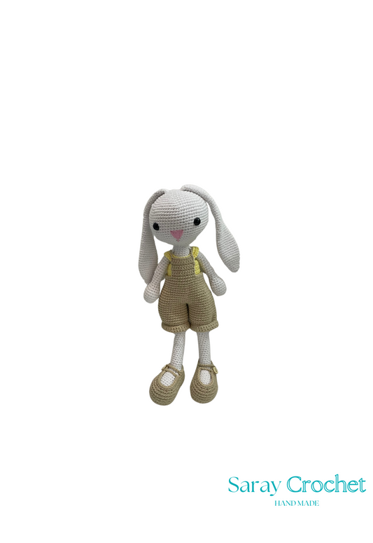 A crochet doll resembling a rabbit with gray body, wearing brown overalls and beige shoes, with the brand name 'Saray Crochet' on the bottom.