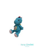 A handcrafted plush toy in the shape of a blue dinosaur with a striped tail and a long striped snout, with the brand name Saray Crochet visible.