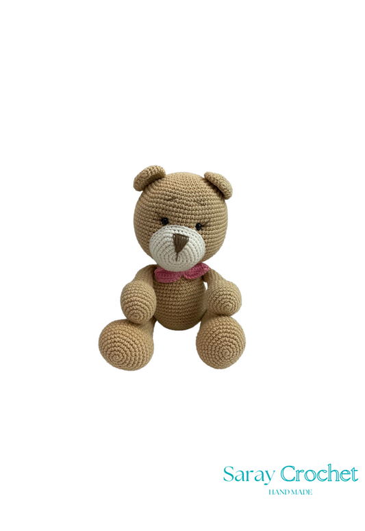 A crochet stuffed toy resembling a bear with a beige body, a pink ribbon around its neck, and a brown nose.