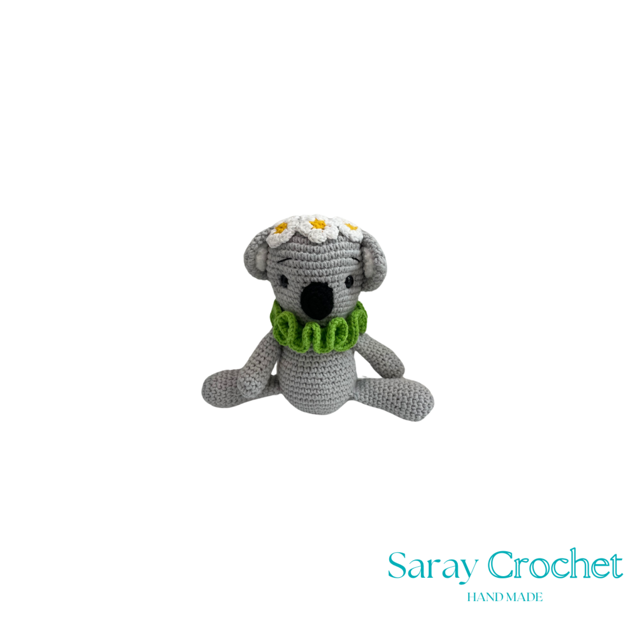 Crocheted koala toy with a green scarf and flower headband on a white background, branded 'Saray Crochet'.