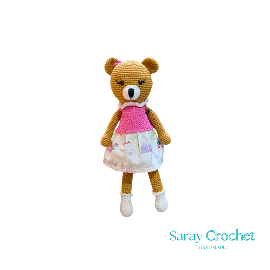 Handmade plush toy resembling a bear with a pink dress and white shoes, with the brand name 'SarayCrochet' visible.