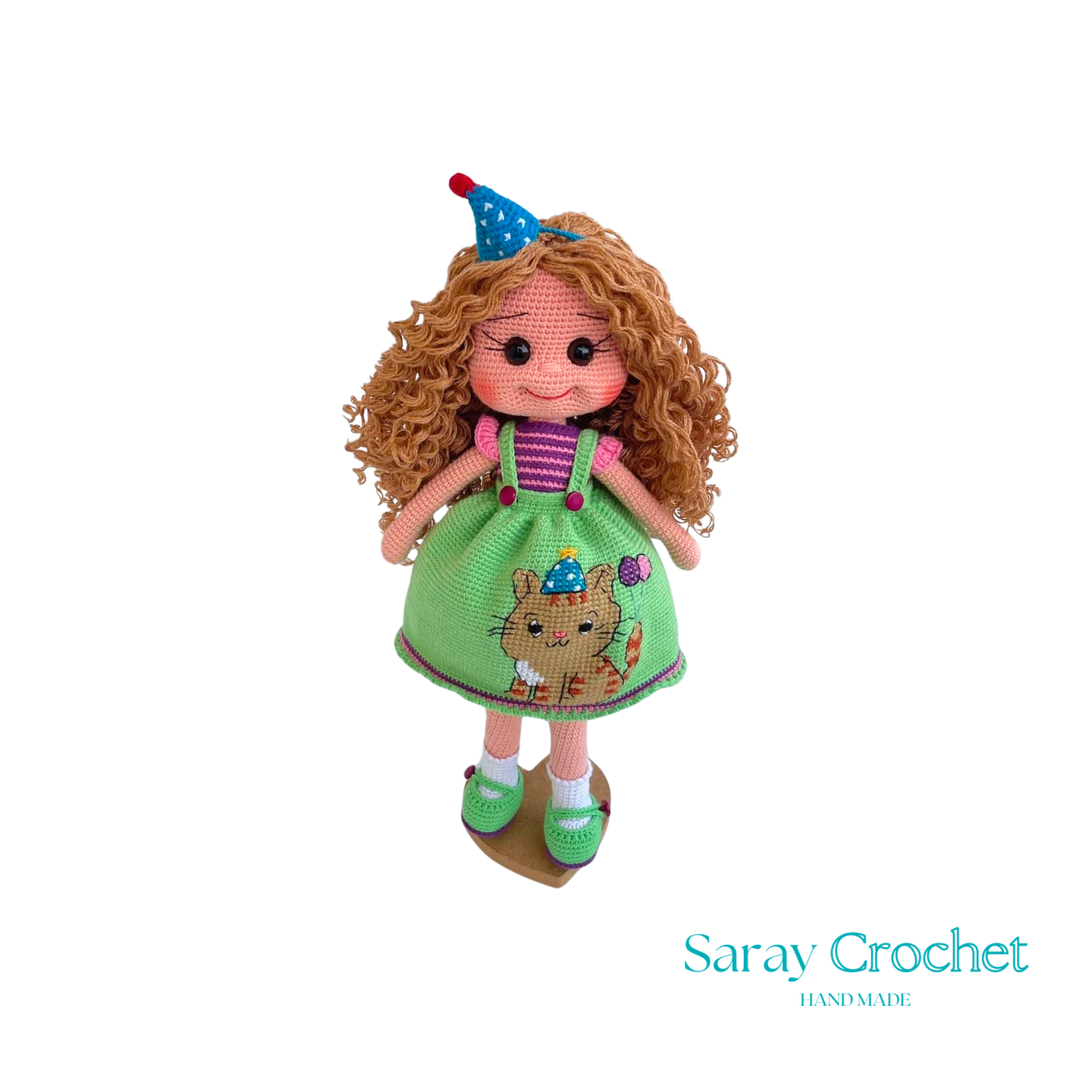 A handcrafted doll with curly orange hair, wearing a green dress with a bear motif and a birthday cap, and green shoes.