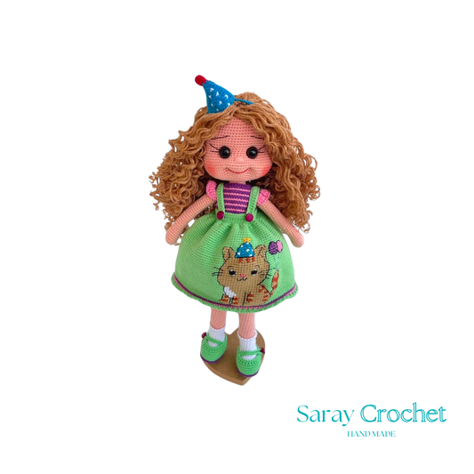 A handcrafted doll with curly orange hair, wearing a green dress with a bear motif and a birthday cap, and green shoes.