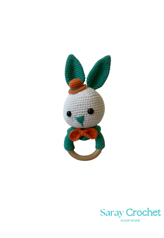 A crochet toy resembling a green and white rabbit with an orange collar and a wooden ring attached to its head.